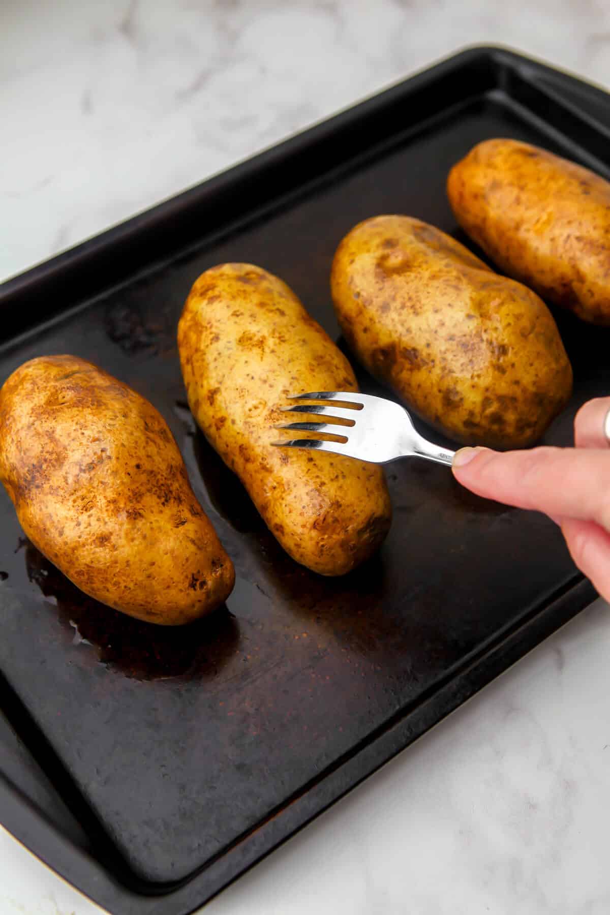 Four potatoes on a baking sheet with someone poking hole in them with a fork.