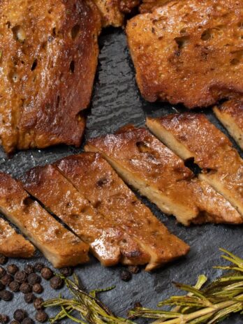 Vegan seitan on a dark cutting board with spices around it.