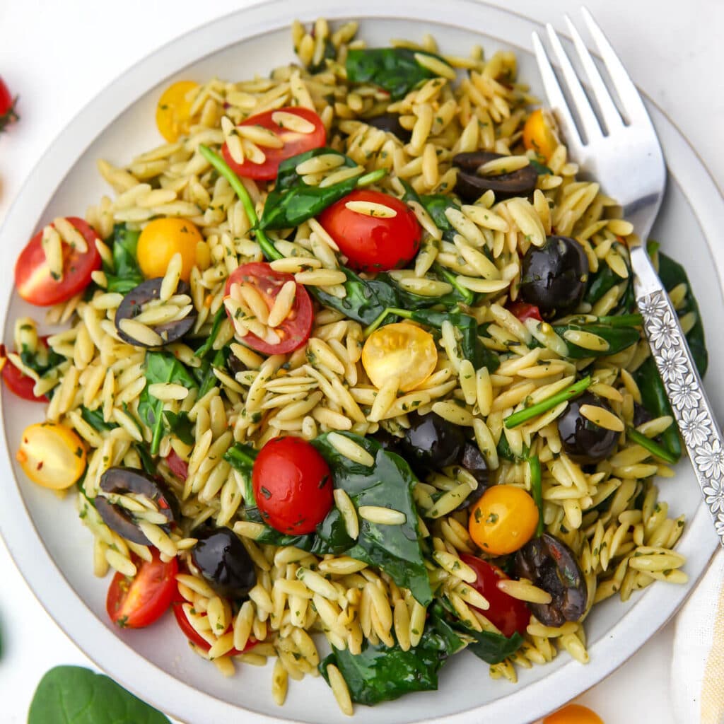 An orzo pesto pasta salad with tomatoes, olives, and spinach on a white plate.