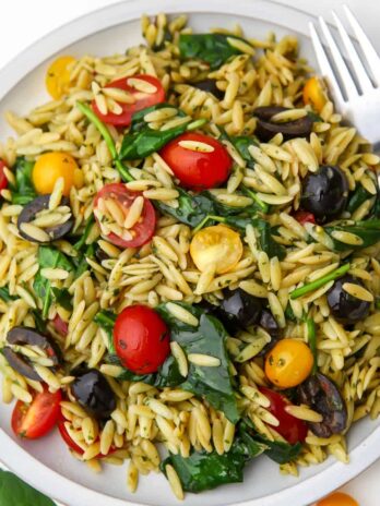 An orzo pesto pasta salad with tomatoes, olives, and spinach on a white plate.