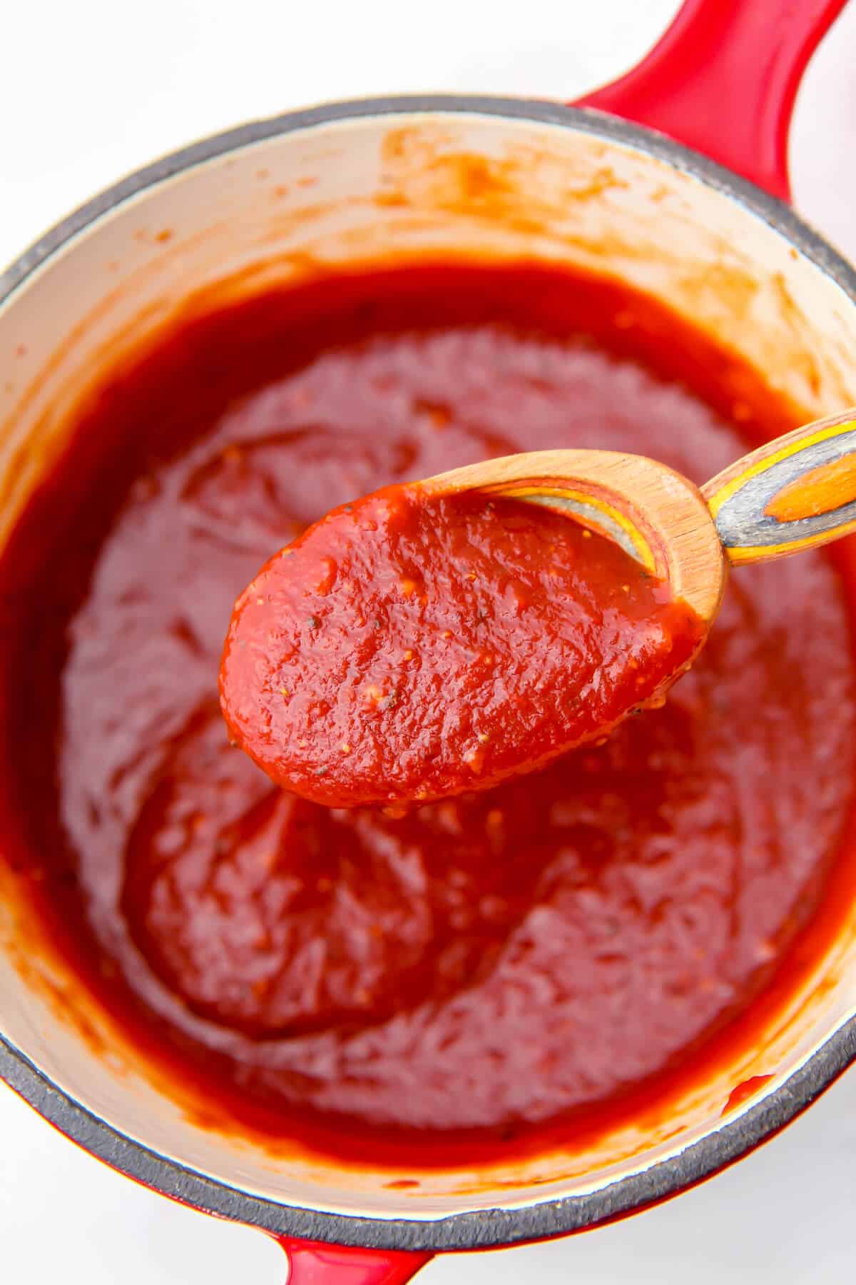 Vegan barbeque sauce being scooped up with a wooden spoon over a red saucepan.