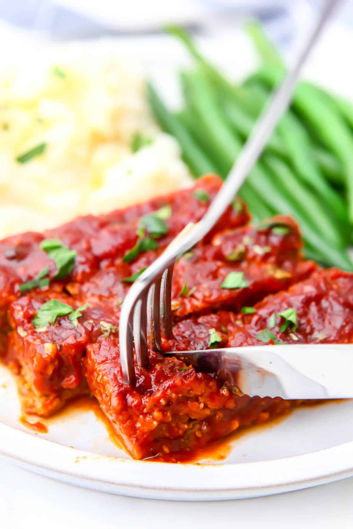 Vegan ribs being cut on a plate with mashed potatoes and green beans.