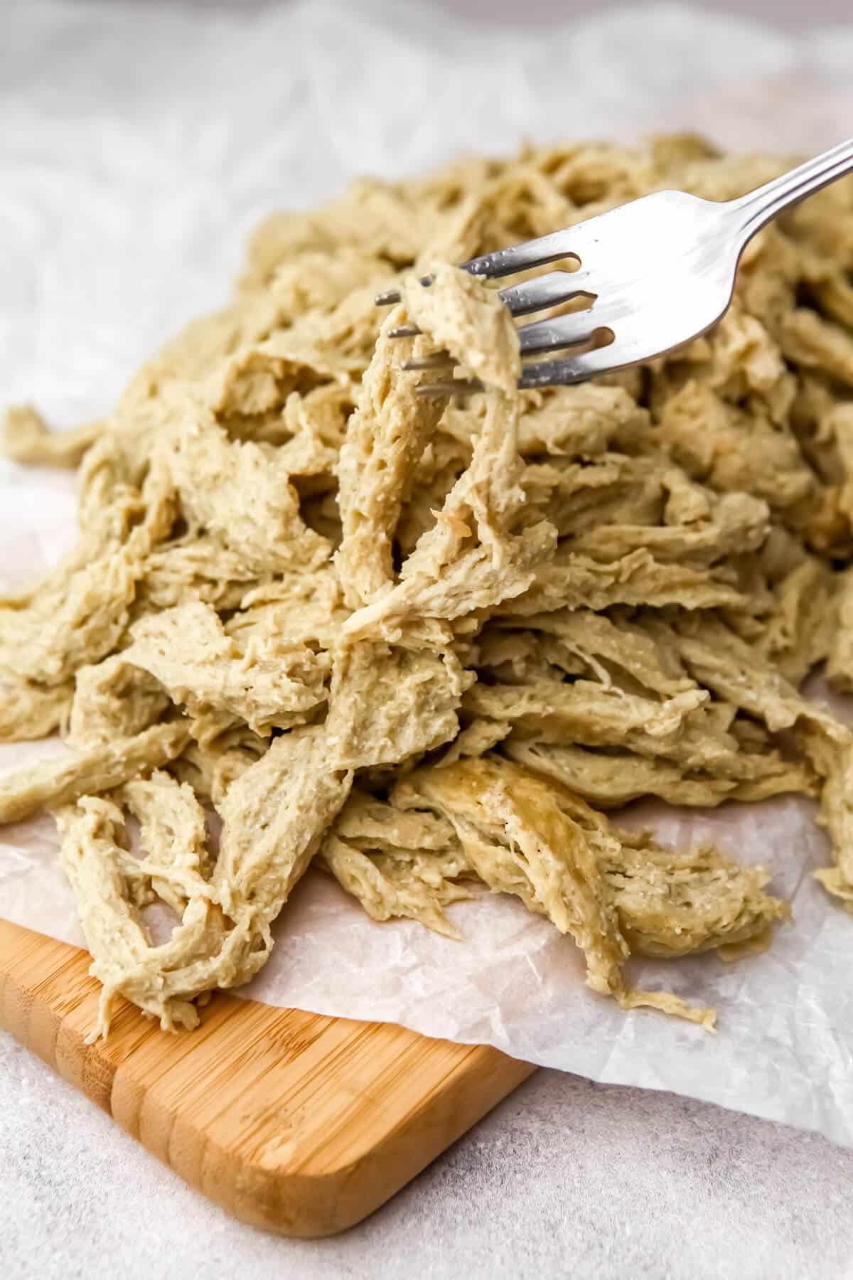 Stringy vegan chicken on a cutting board with a fork holding a piece of the faux chicken up.