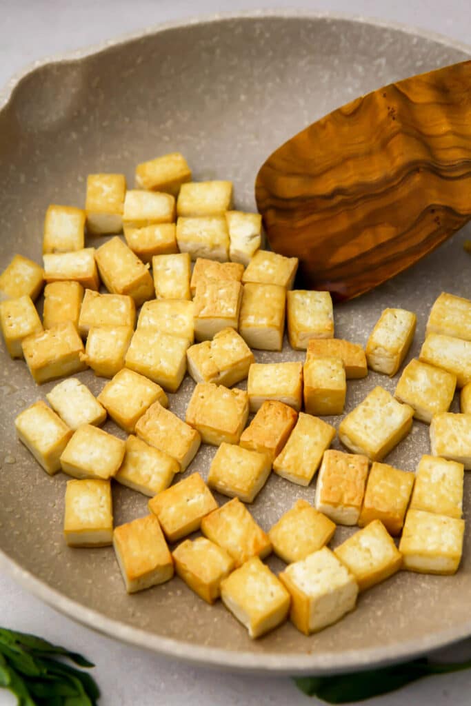 Cubes of tofu pan-frying until golden brown in a white frying pan.