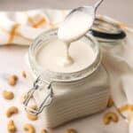 A glass container with a lid filled with vegan heavy cream made from cashews with a spoonful dripping cream into the jar.