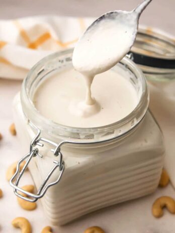 A glass container with a lid filled with vegan heavy cream made from cashews with a spoonful dripping cream into the jar.