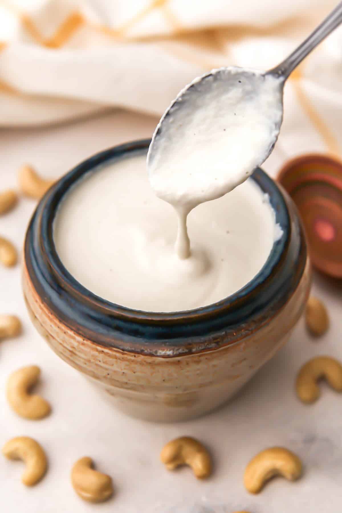 A jar of cashew cream with a spoon over it and cashews around it.