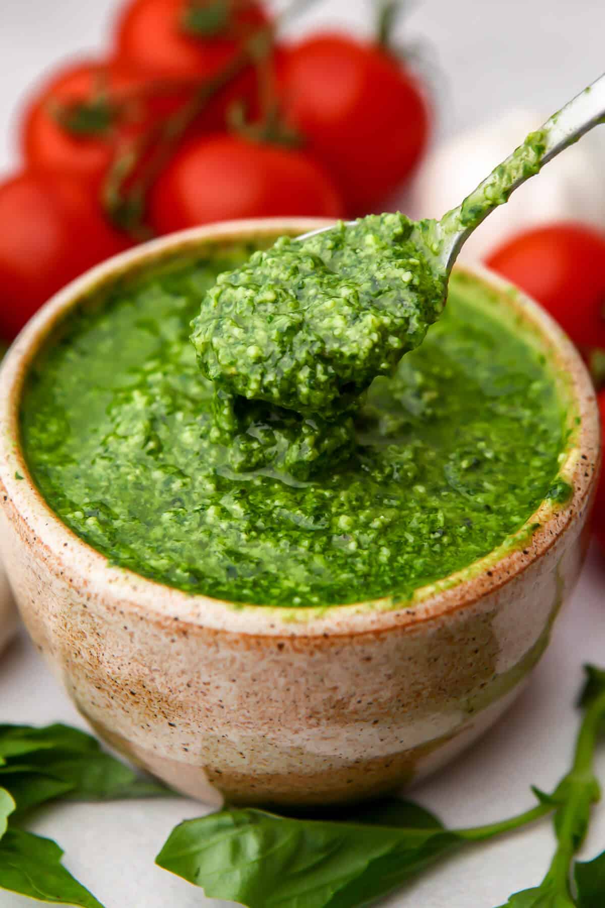 A bowl of vegan pesto with tomatoes and garlic behind it and basil leaves around it.