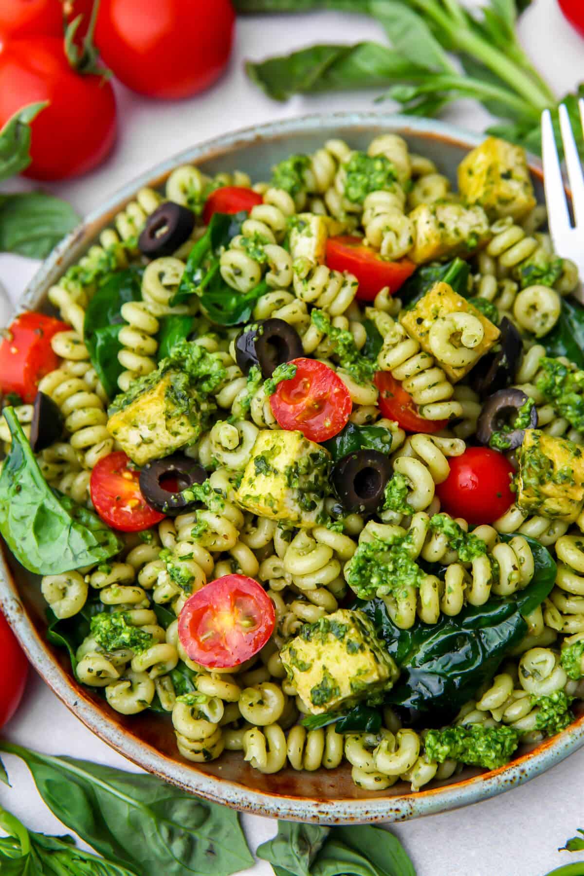 A plate full of vegan pesto pasta with cherry tomatoes, black olives, spinach, and tofu.
