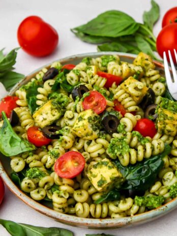A plate filled with vegan pesto pasta with fresh basil, tomatoes, and garlic on the side.