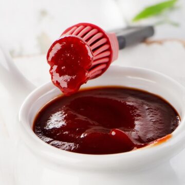 A white bowl filled with BBQ sauce with a silicon brush resting on it.