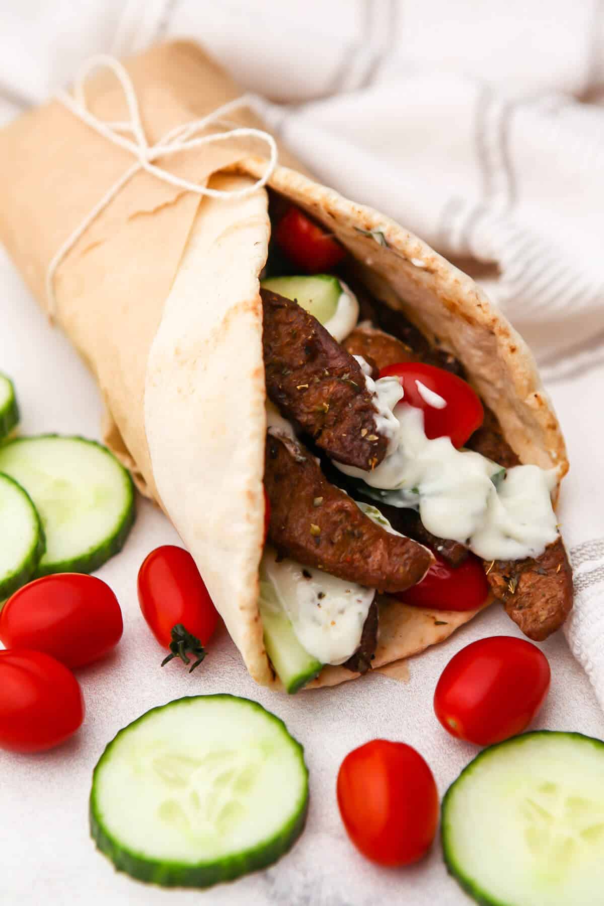 A seitan gyro wrapped in parchment paper with cherry tomatoes and cucumbers around it.