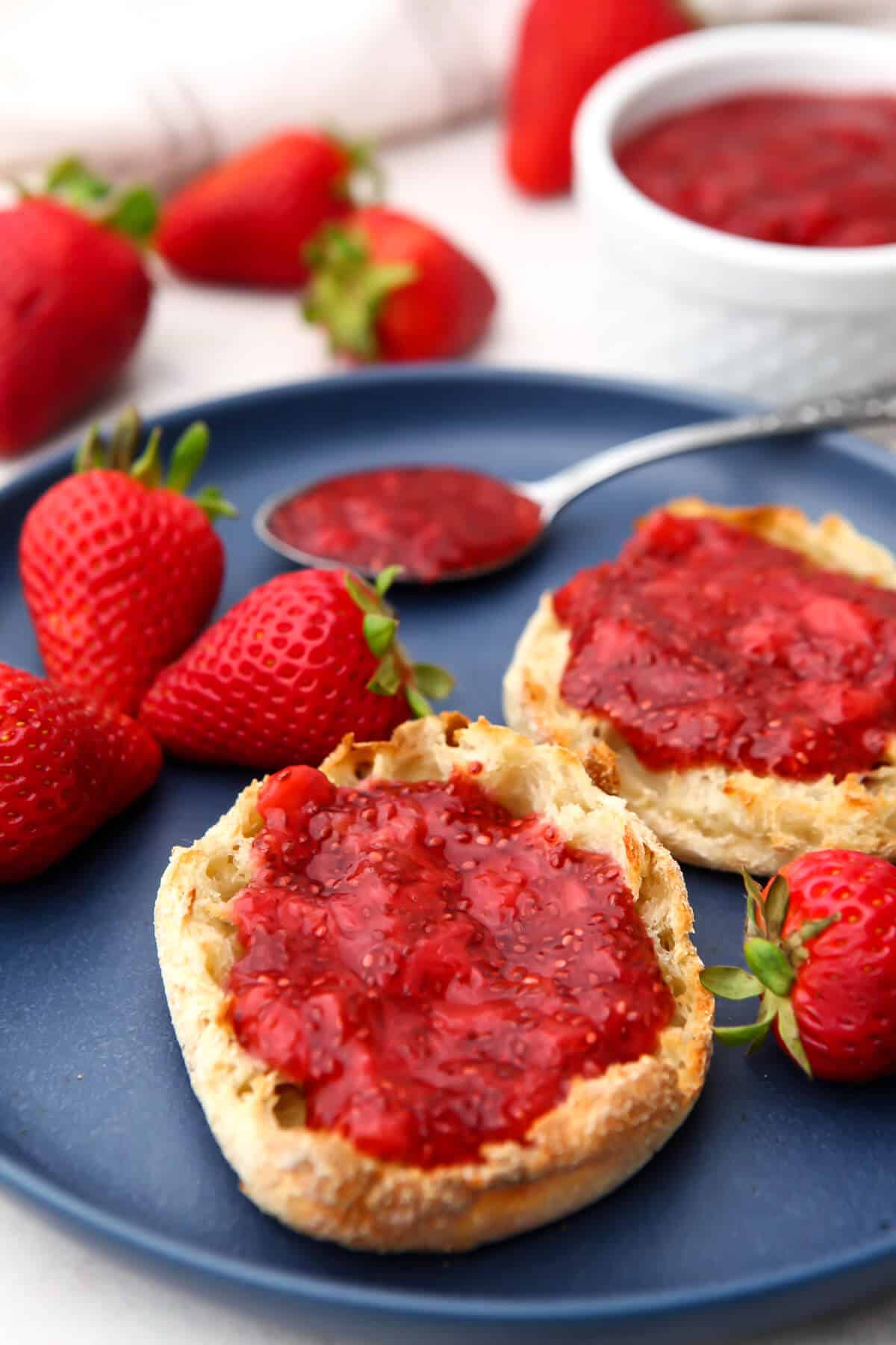 Strawberry chia seed jamb spread on two English muffins on a blue plate with strawberries around it.