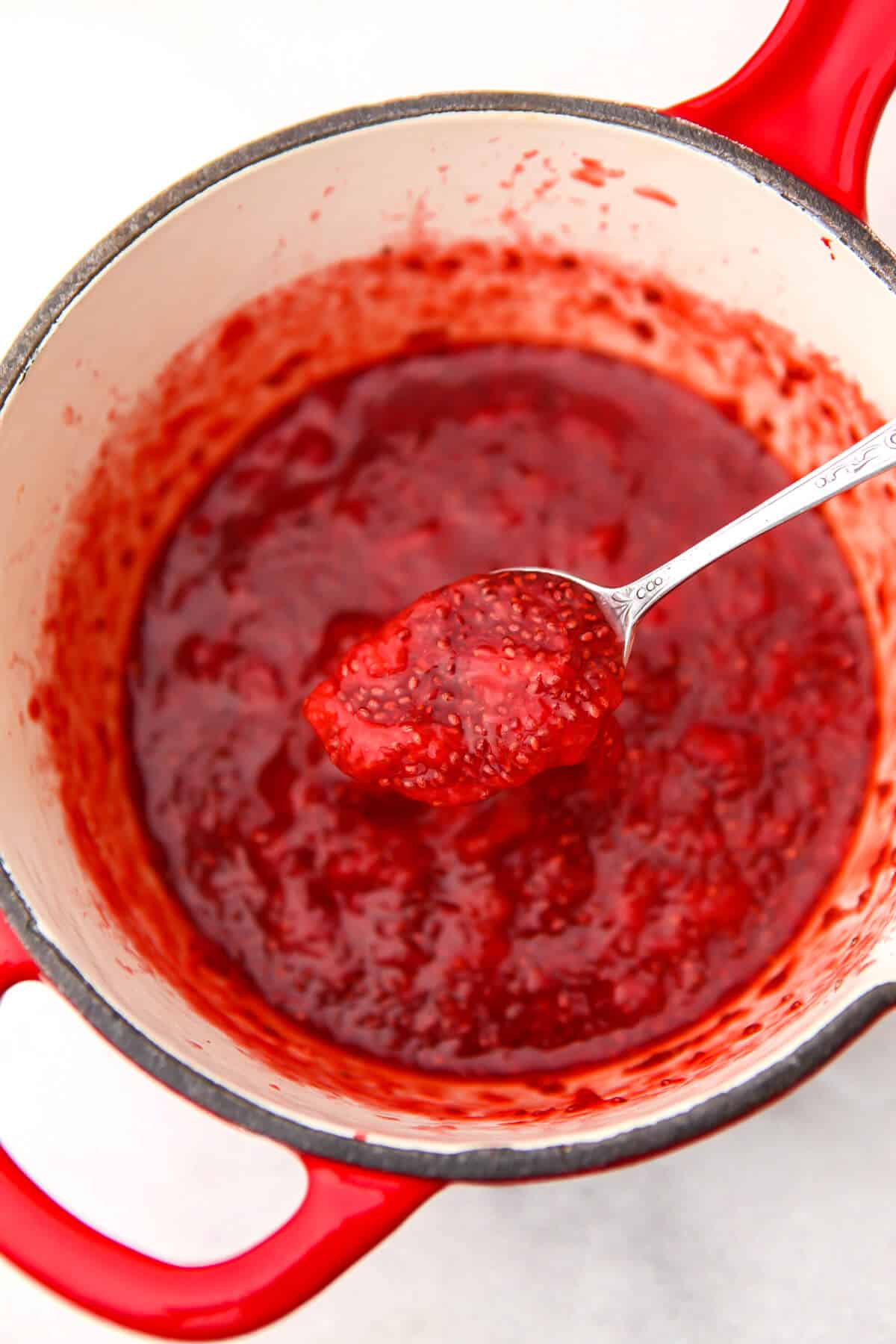 A red saucepan filled with fresh strawberry jam made with chia seeds being scooped up with a spoon.