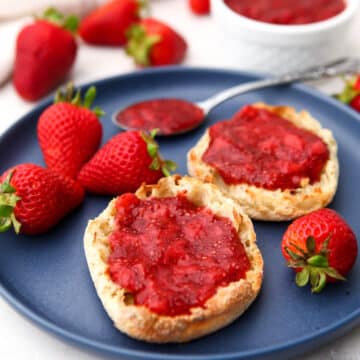 Strawberry chia seed jamb spread on two English muffins on a blue plate with strawberries around it and a white bowl of jam behind it.