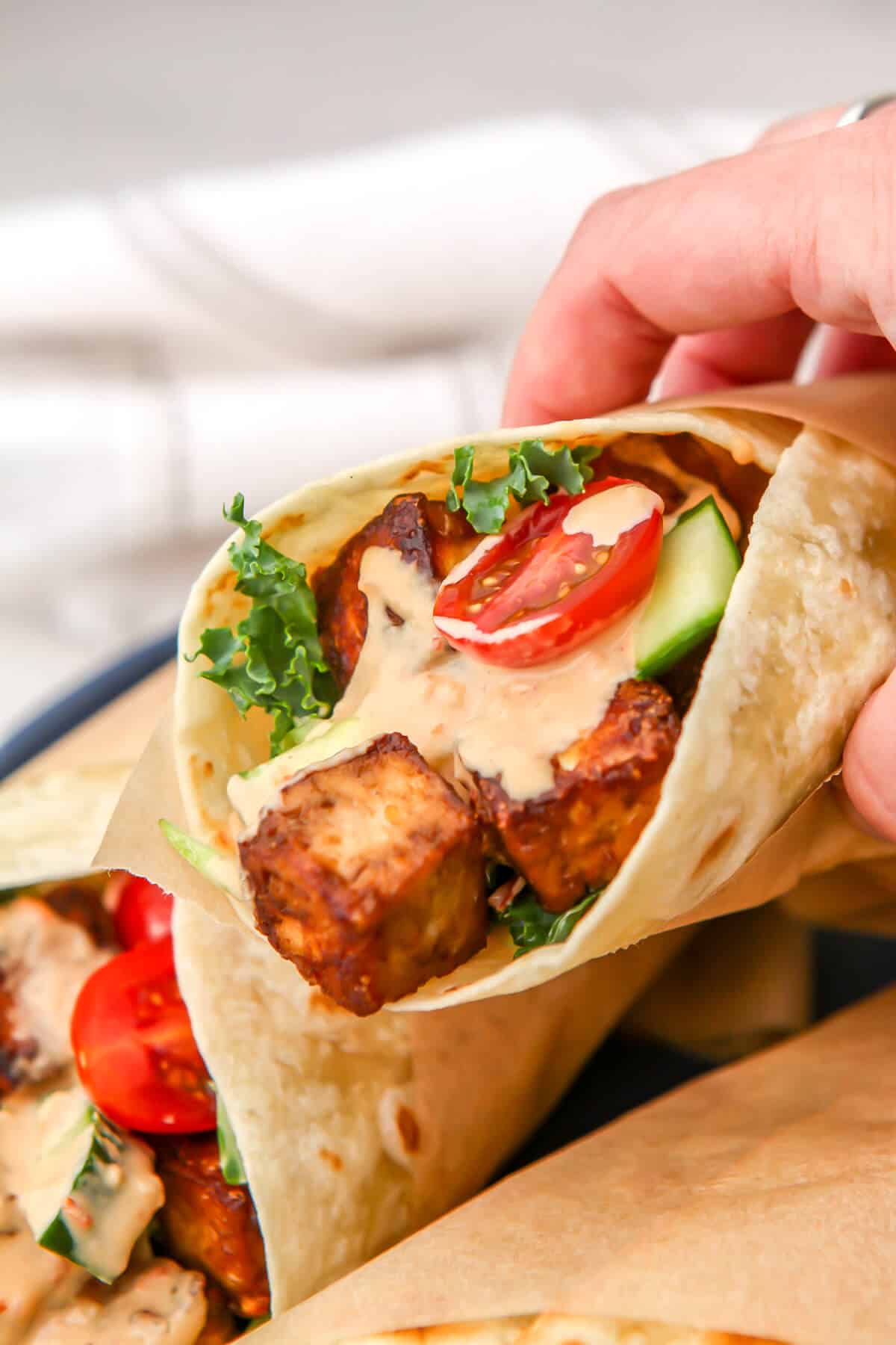 Someone holding a tofu wrap with veggies and sauce on it.