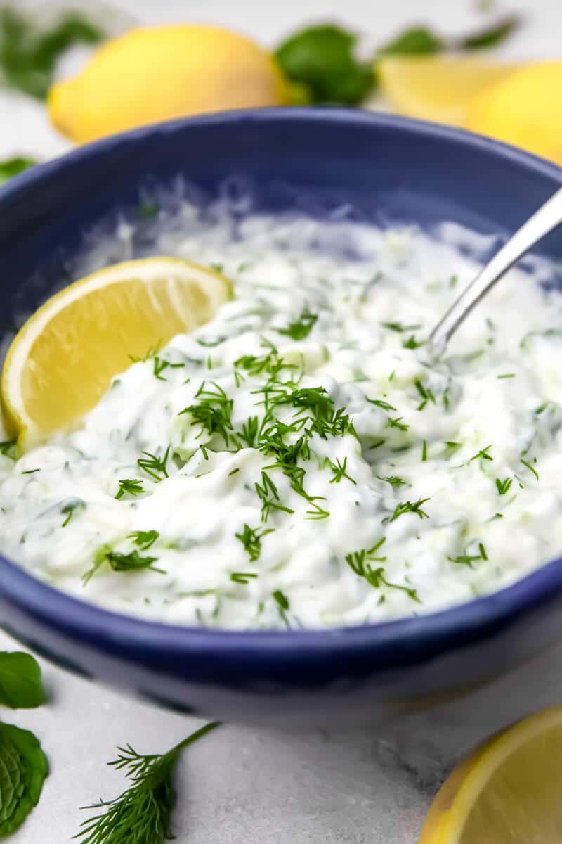 Abowl of vegan tzatziki sauce in a blue bowl with lemon and herbs around it.