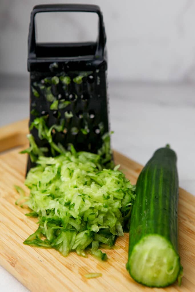 An English cucumber shredded on a cutting board with a grater behind it.