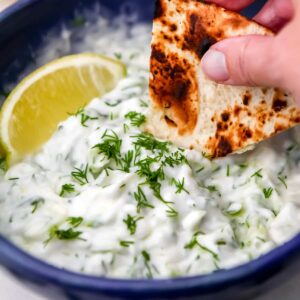 Someone dipping a pita bread triangle into a bowl of vegan tzatziki sauce.