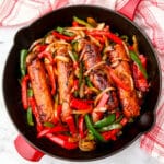 A top view of a red iron skillet with vegan sausages cooked with onions and peppers.