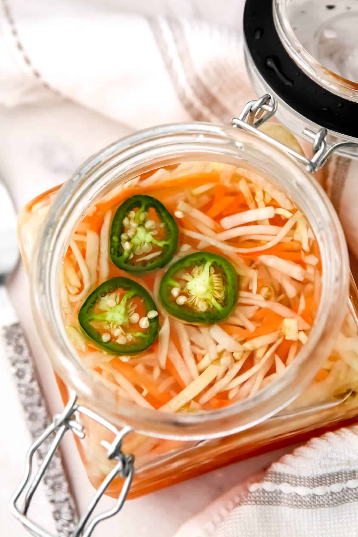 A glass jar filled with pickled carrots and daikon with jalapeno slices on top.,