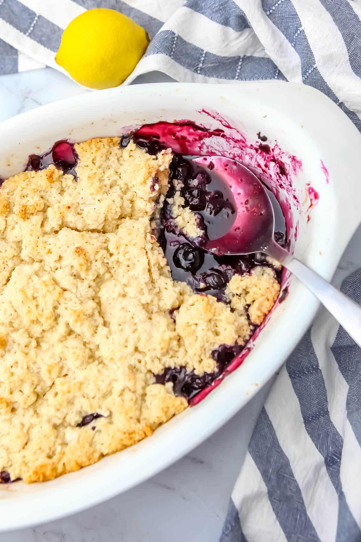 A vegan blueberry cobbler in a white baking dish with a blue and white tea towel and a lemon on the side.