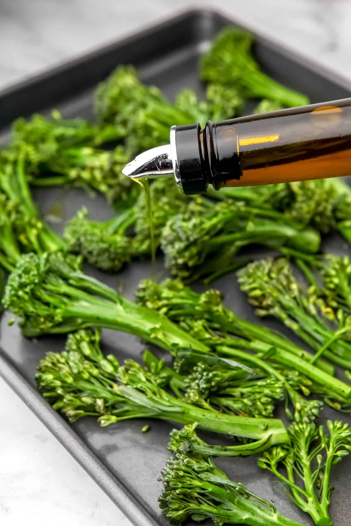 Raw tendersteam broccoli on a cookie sheet being drizzled with olive oil.