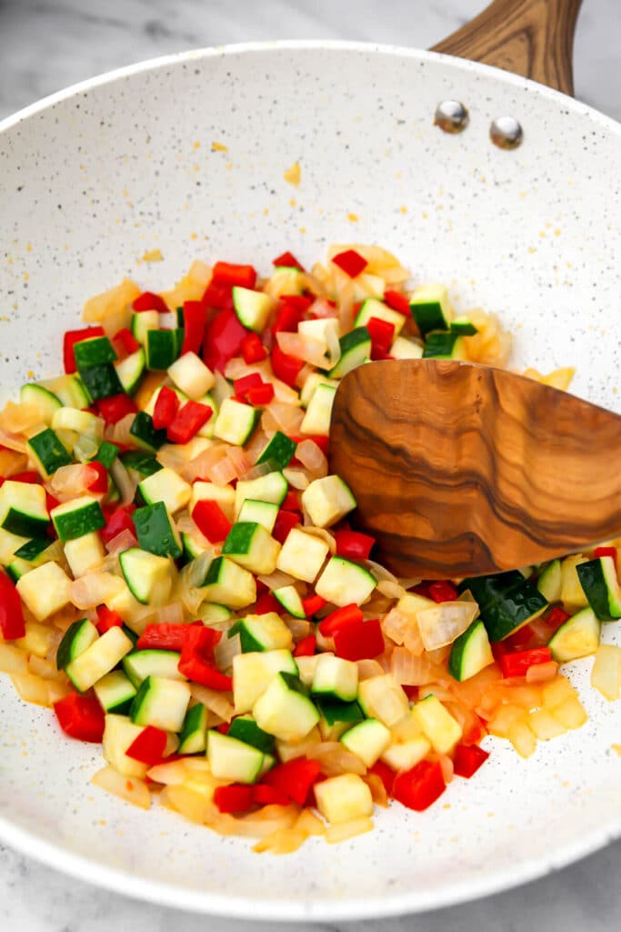 Onions, garlic, zucchini, and red bell pepper frying in a large white wok.