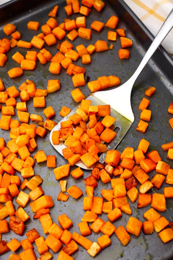 Small cubes of butternut squash roasting on a baking sheet being tossed with a spatula.