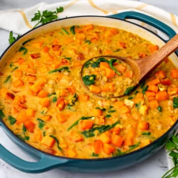 A large blue skillet full of Butternut squash and chickpea curry with red lentils, spinach, and coconut milk with a wooden ladle in it.