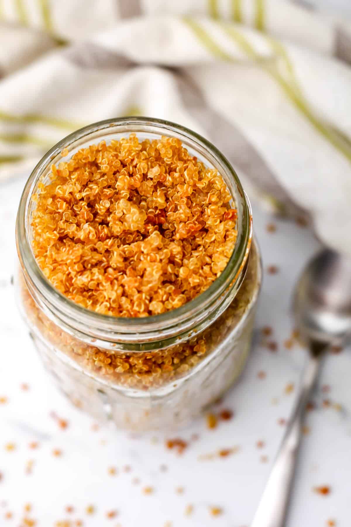 A top view of a jar filled with crunchy quinoa with a spoon on the side and a tea towel behind it.