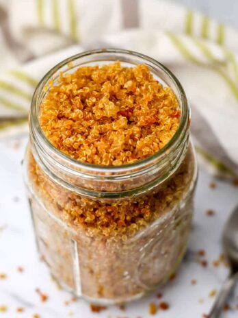 A glass jar full of toasted crunchy quinoa with a spoon and a tea towel on the side.