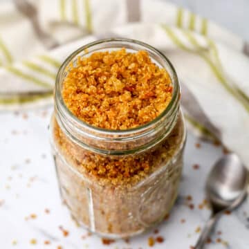 A glass jar full of toasted crunchy quinoa with a spoon and a tea towel on the side.
