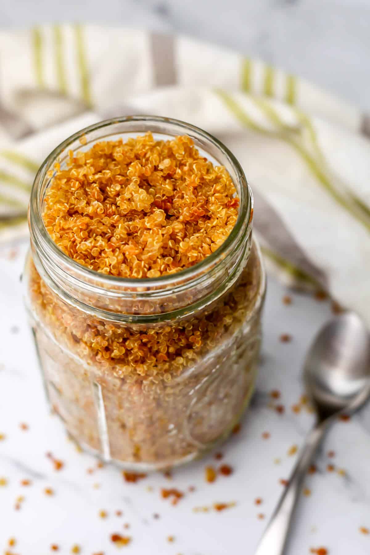 A glass jar full of crunchy quinoa with a spoon on the side and a tea towel behind it.