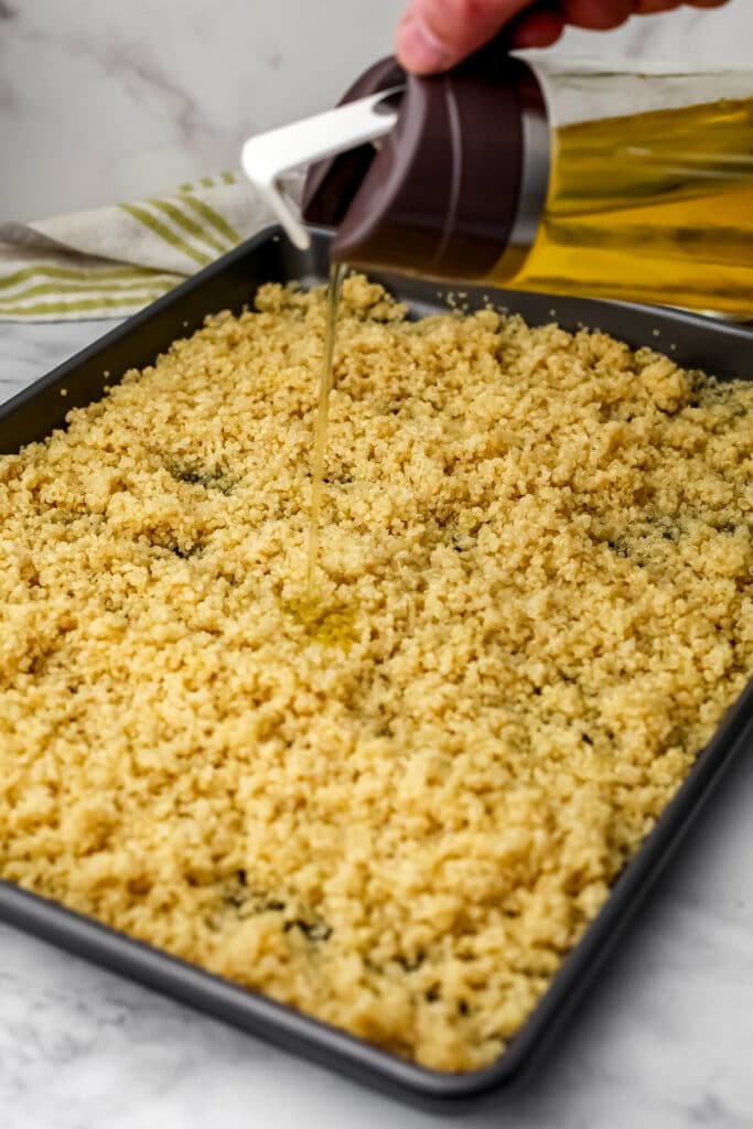 Cooked Quinoa on a baking sheet being drizzled with some olive oil before baking to make crispy quinoa.
