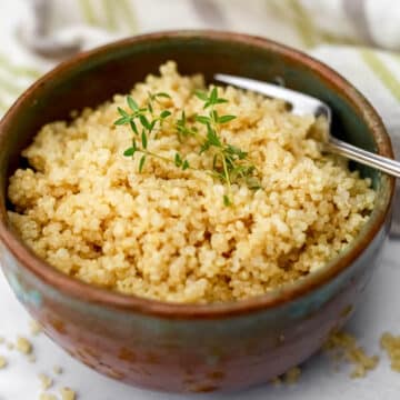A bowl of fluffy quinoa in a brown bowl with a sprig of some herbs on top.