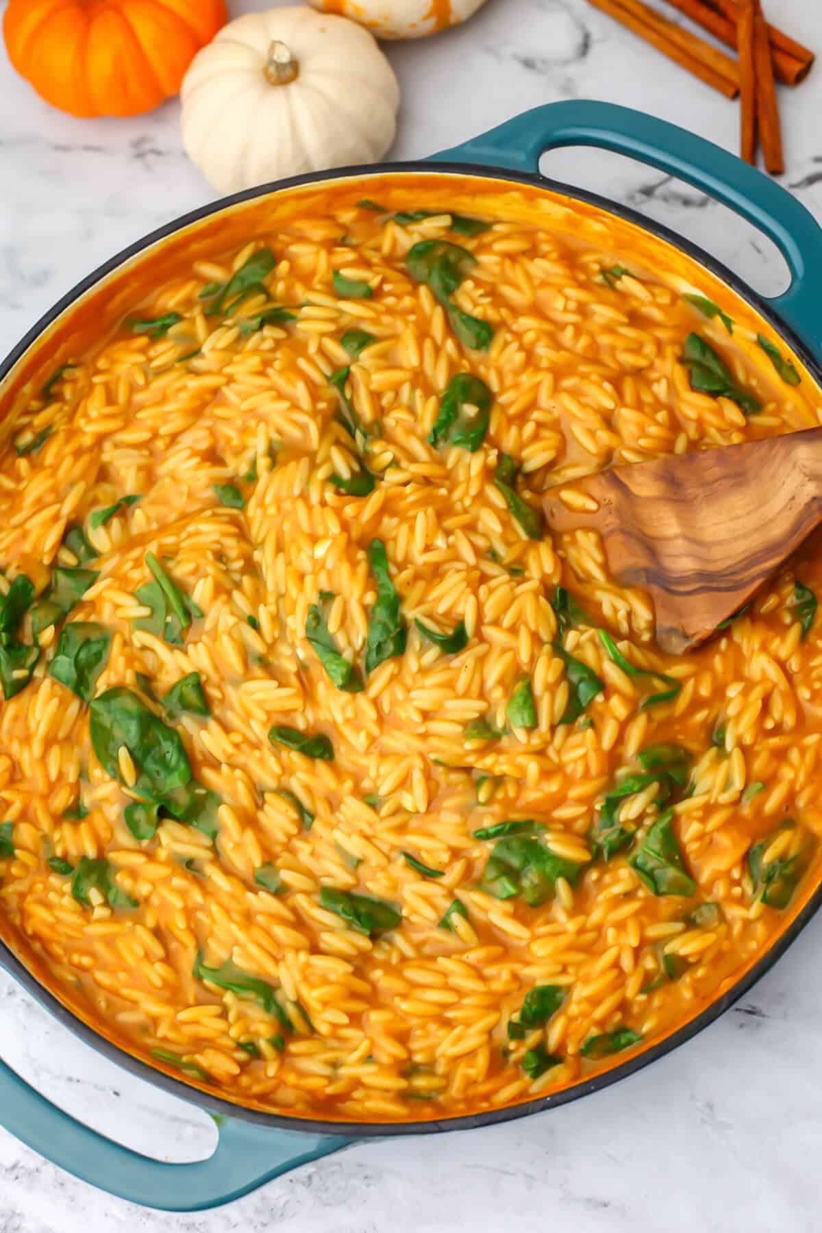 A top view of a vegan pumpkin orzo with spinach dish in a blue skillet with little pumpkins around it.