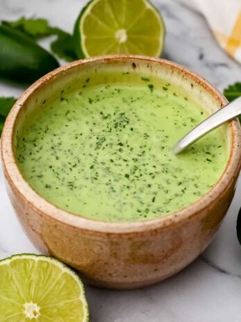 A bowl of vegan cilantro lime crema with limes, jalapenos, and cilantro on the side.