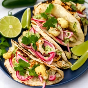 A blue plate with three cauliflower tacos on it with pickled onions and cilantro sauce with lime wedges on the side.