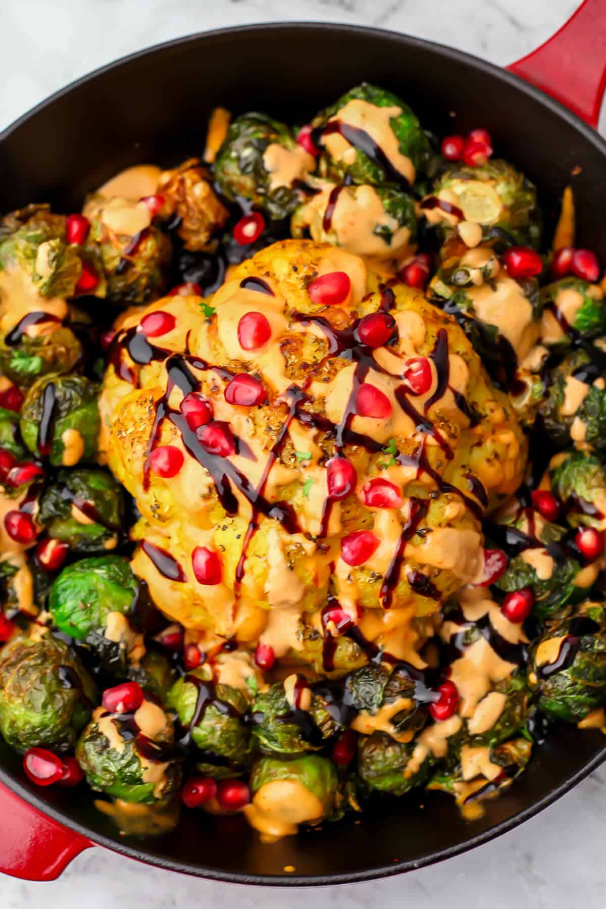 A top view of a whole head of cauliflower that has been roasted and drizzled with spicy tahini sauce, balsamic glaze and pomegranate seeds.