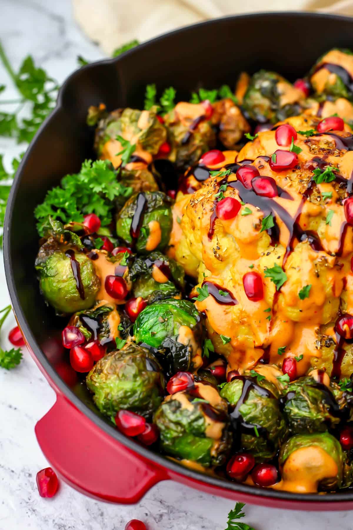 A whole roasted head of cauliflower in a red iron skillet with brussels sprouts around it and drizzled with tahini sauce.