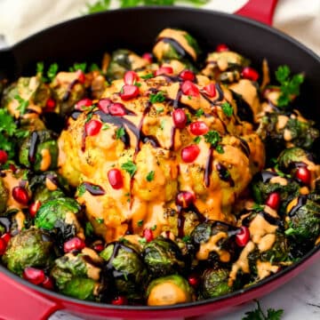 A whole roasted cauliflower head with brussels sprouts around it and drizzled with tahini dressing, balsamic glaze and sprinkled with pomegranate seeds.