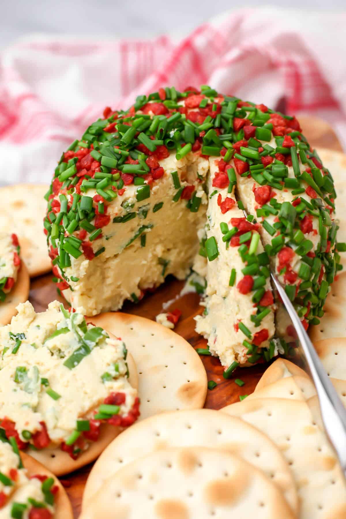 A vegan cheese ball coated in chopped chives and vegan bacon bits with crackers around it.