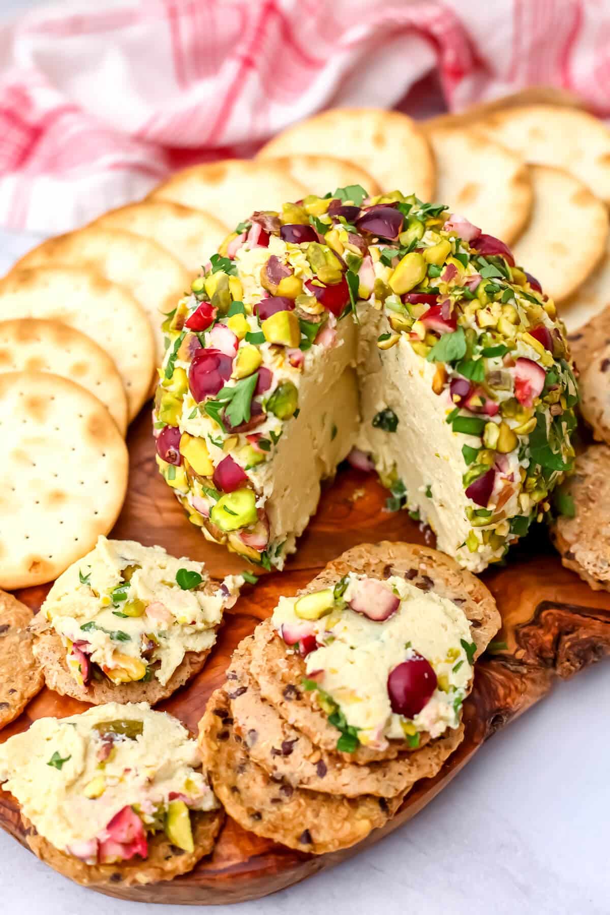 A vegan tofu cheese ball rolled in pistachios and cranberries on a cutting board with crackers.