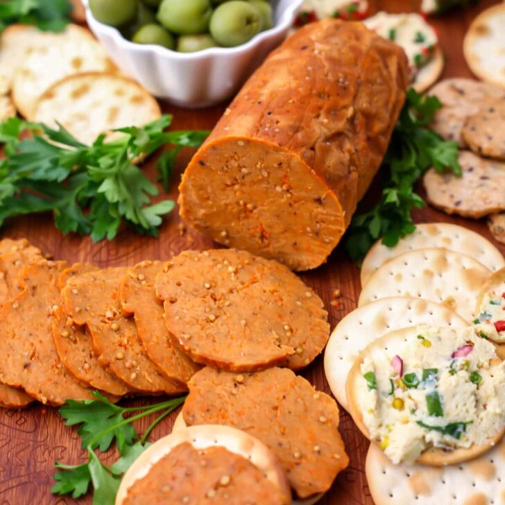 A vegan summer sausage made from seitan sliced on a wooden cutting board with crackers on the side.