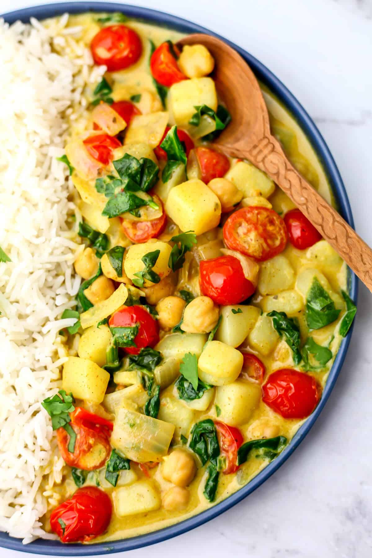 A top view of a plate of vegan potato curry with Balti seasoning with chickpeas, spinach, and tomatoes with rice on the side.