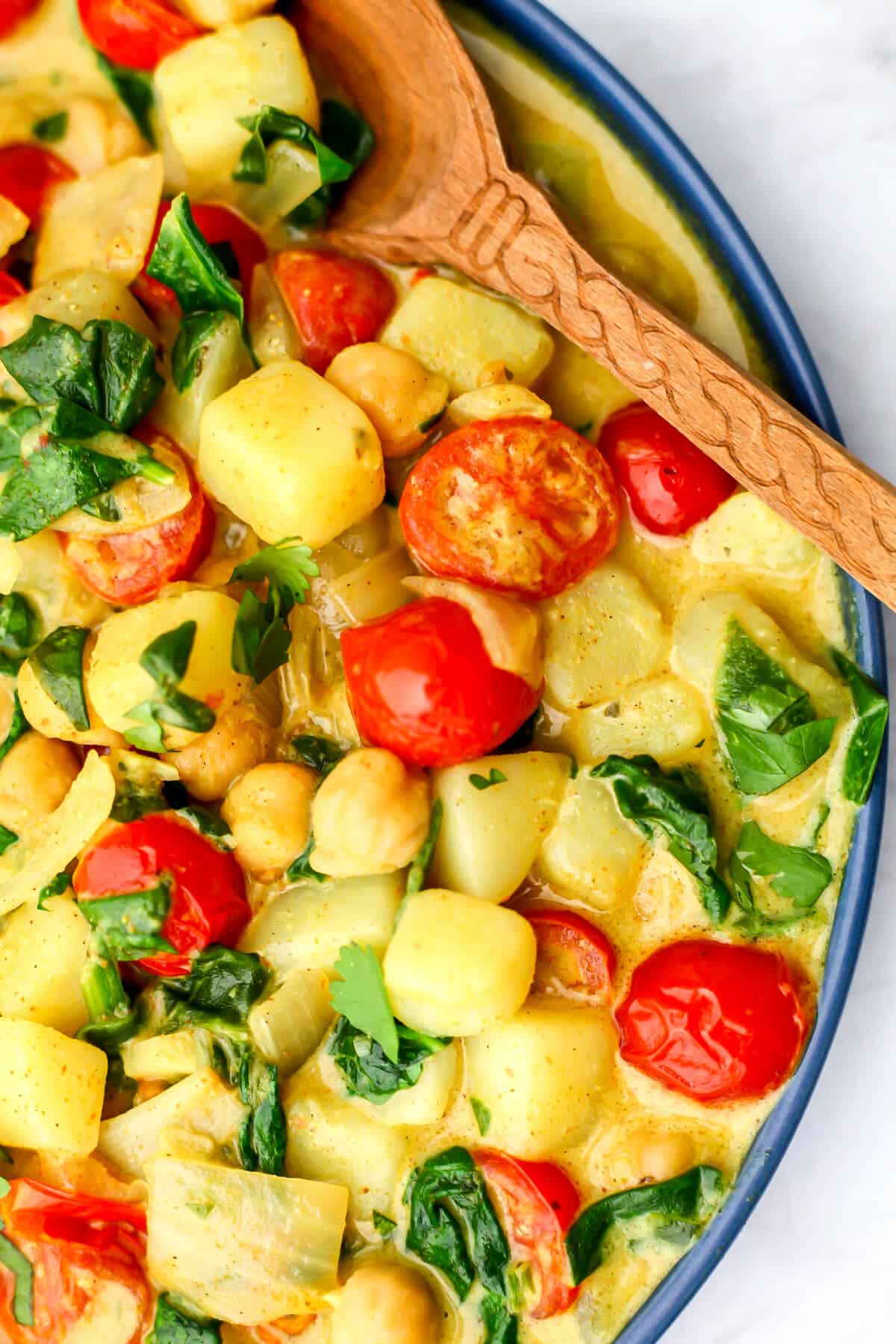 A blue plate filled with potato chickpea curry with spinach and tomatoes with a wooden spoon on the side.