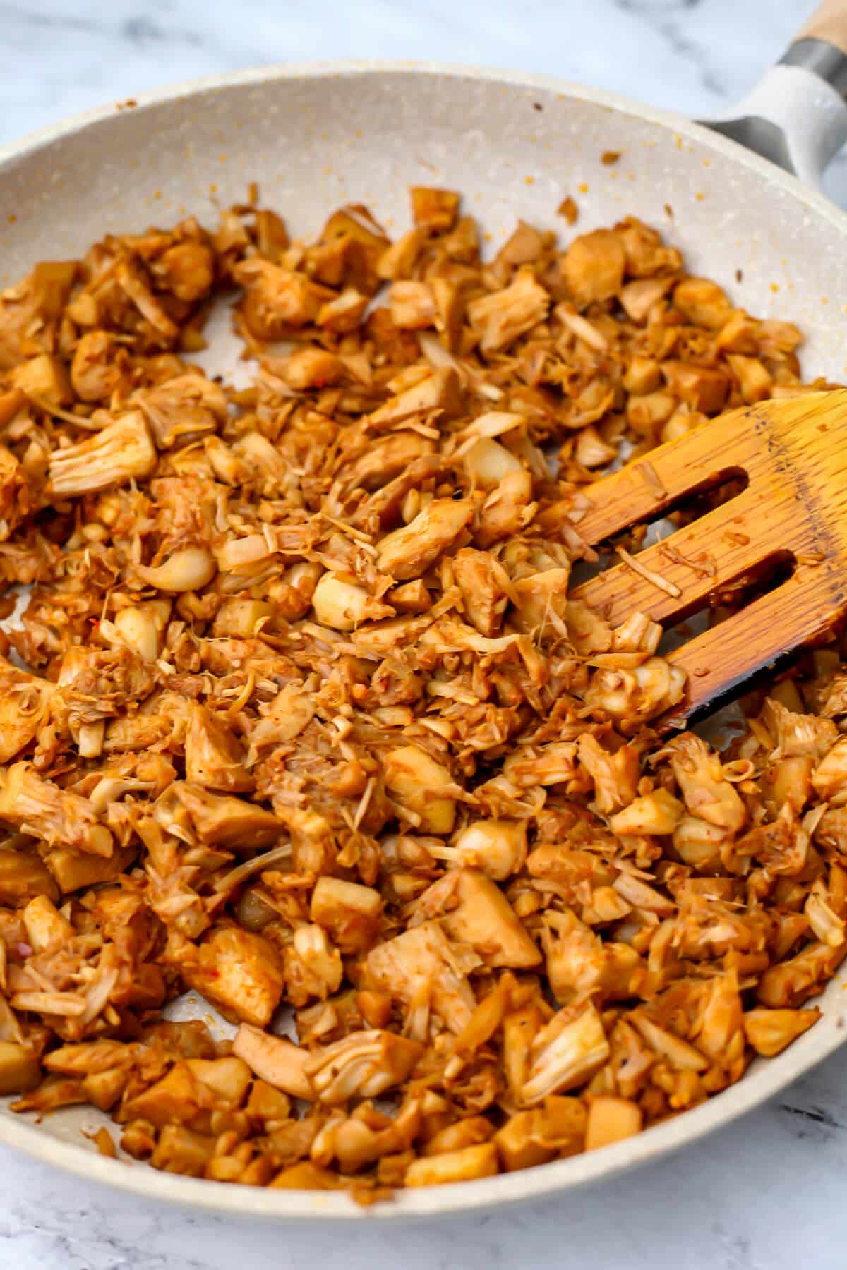 Seasoned jackfruit sautéing in a frying pan being stirred with a wooden spatula.