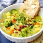 A white bowl filled with creamy vegan broccoli and potato soup with a piece of bread dipped in it.