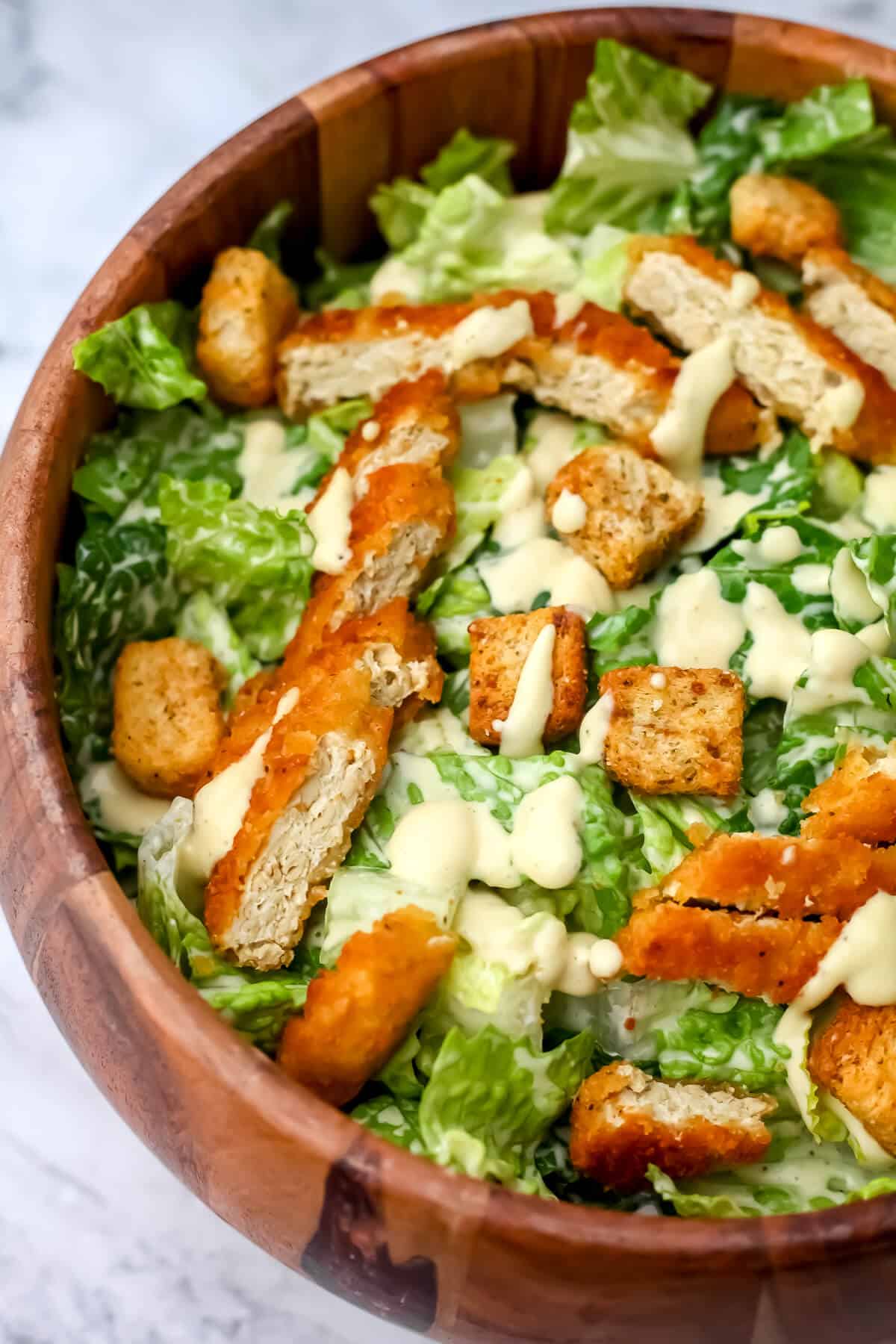 A top view of a wooded salad bowl filled with vegan chicken Caesar salad with dressing, faux chicken, and croutons on top.